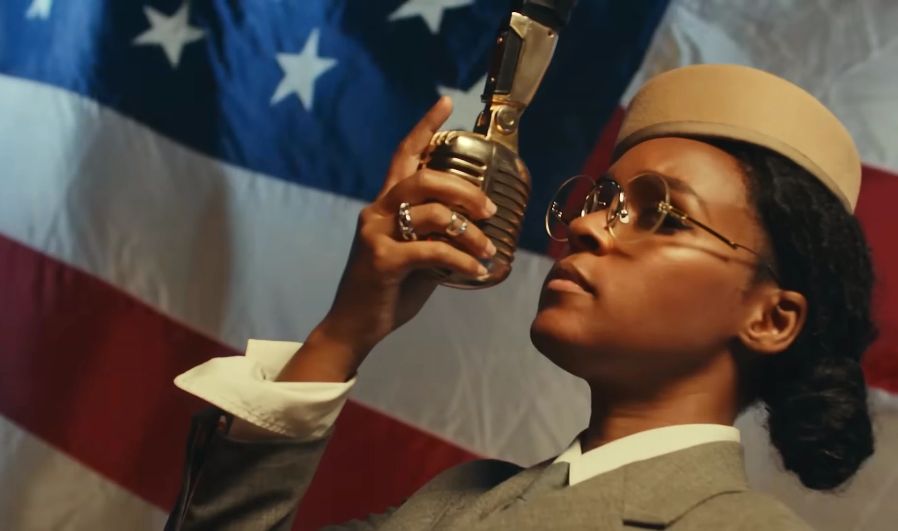 Janelle Monáe. Turntable clip. vestida de militar sujeta un micrófono vintage, el fondo de la imagen es la bandera de los EEUU.feedcult.com.webp