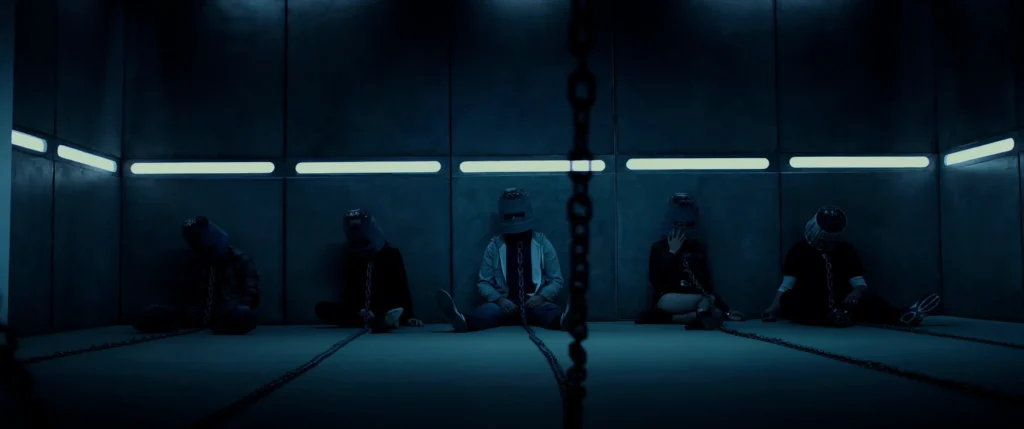Jigsaw (2017) víctimas sentadas en el suelo de la sala, con el casco de acero y encadenados. El juego está a punto de comenzar.
